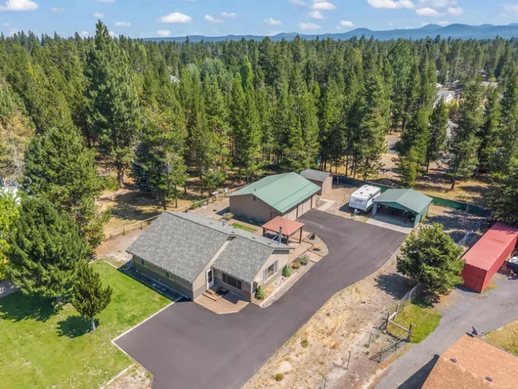 Aerial view of a single-story tan home with a grey roof, a long paved driveway, a detached green-roofed garage and tall pines at 55872 Wood Duck Dr, Bend, OR 97707